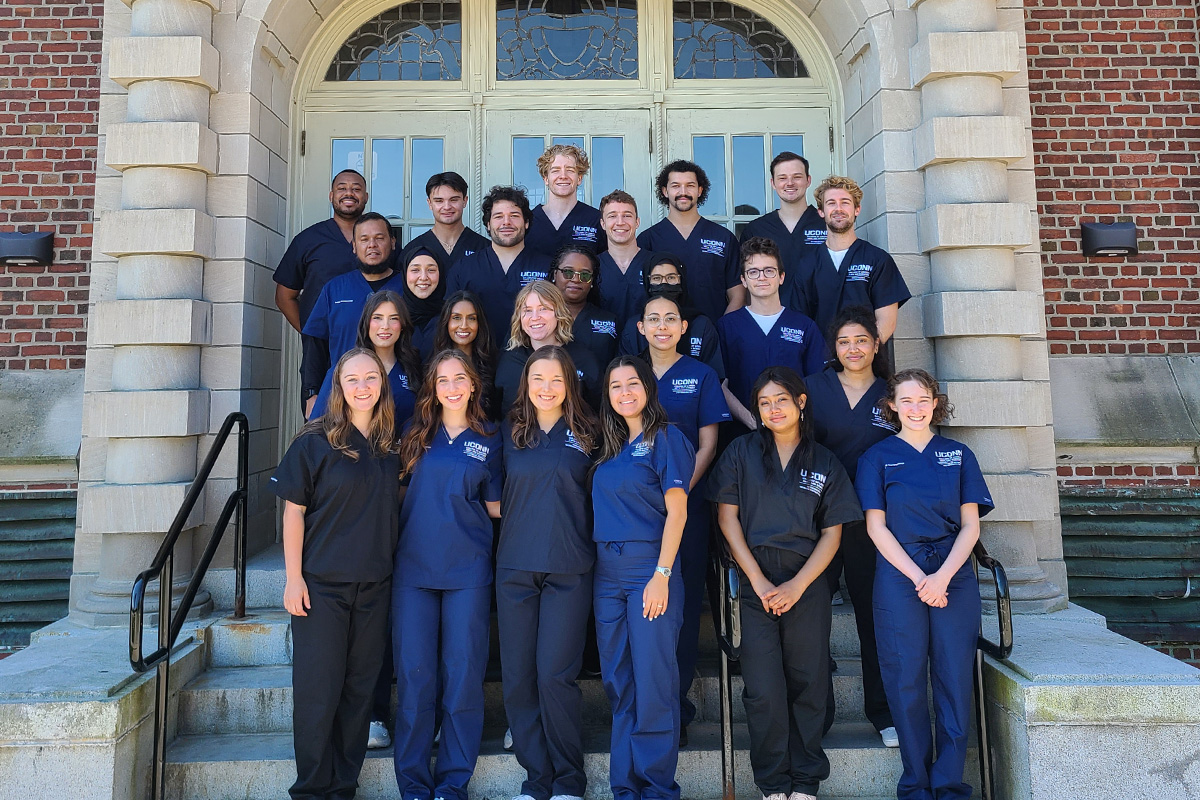 The 2024-2025 class poses in front of Beach Hall wearing surgical scrubs. 