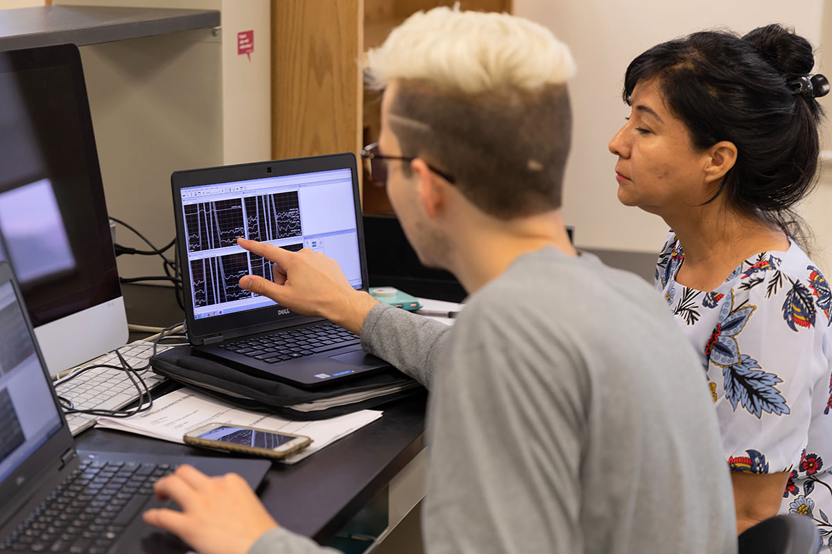 Two students in a research lab review data on a laptop, with one pointing to the screen.